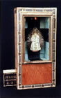 Eine Heiligenfigur ist hinter Glas in eine Zigarrenkiste montiert. Durch Einwurf einer Münze geht ein Licht an und eine Fahne springt heraus.